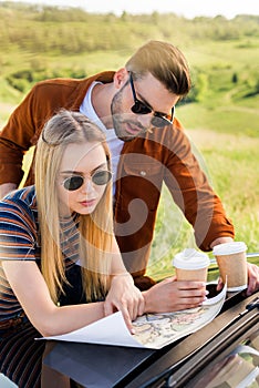stylish couple of tourists with paper cups of coffee looking for destination on map near car in rural