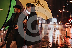 Stylish couple using an ATM machine on a rainy day. They are withdrawing money while having fun conversation and smiling