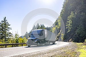 Stylish dark gray big rig semi truck transporting cargo in dry van semi trailer running on the winding mountain road