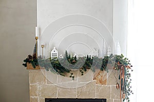 Stylish decorated christmas fireplace. Modern christmas trees, houses, pine cones and spruce branches on fireplace mantel with