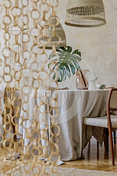 Stylish dining room interior with wooden table, design chairs, rattan pendant lamp, tropical leaf in vase, beautiful plates.