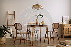 The stylish dining room with round table, rattan chair, wooden commode, pock up poster and kitchen accessories. Beige wall with