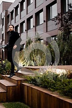 Stylish dressed businessman is walking down by stairs, looking at the side, posing for photo outdoor. Successful