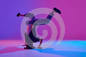Stylish dressed young dancer, guy performing breakdance tricks in mixed neon light against vibrant gradient background.