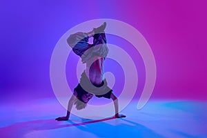 Stylish dressed young dancer, guy performing headstand in motion in mixed neon light against vibrant gradient background