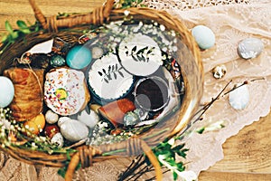 Stylish Easter basket with modern eggs, easter bread cake, ham, beets, sausage, butter and green branches on rustic fabric with