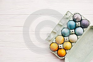 Stylish easter eggs in carton tray on white wooden background, top view. Modern colorful easter eggs painted with natural dye in