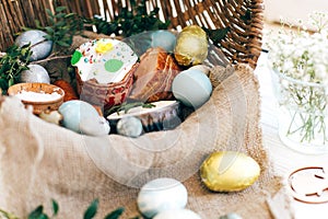 Stylish Easter eggs, easter bread cake, ham, beets, sausage, butter, green branches in wicker basket on white wooden background