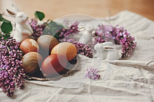 Stylish Easter eggs on rustic plate, white bunnies and lilac flowers on linen rural fabric. Natural dyed easter eggs and spring