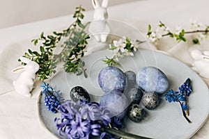 Stylish easter eggs on vintage plate, bunny and spring flowers on rustic table. Happy Easter! Natural dye blue eggs, purple