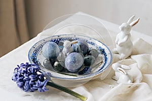 Stylish easter eggs on vintage plate, bunny and spring flowers on rustic table. Happy Easter! Natural dye blue eggs, purple