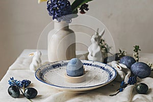 Stylish easter table setting. Easter egg in bunny figurine on vintage plate and spring flowers on rustic table. Happy Easter!