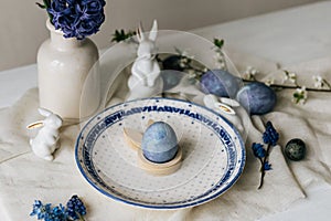 Stylish easter table setting. Easter egg in bunny figurine on vintage plate and spring flowers on rustic table. Happy Easter!