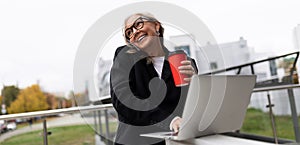 Stylish elderly business woman works on a laptop online on the terrace of an office building and talks on a mobile phone