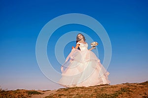 Stylish, elegant bride in a white dress stands on a mountain and poses on a background of blue sky, sunset