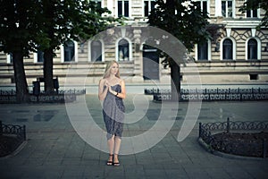 A stylish elegant girl in a dark dress with blond hair stands beautifully in a park alley on a city street