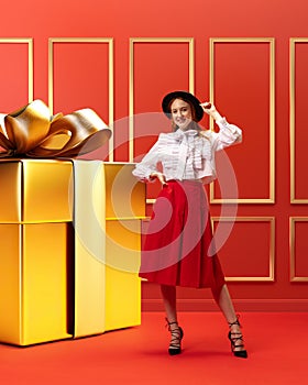 Stylish, elegant young girl standing near giant golden present box, Christmas gift against red background. Happiness