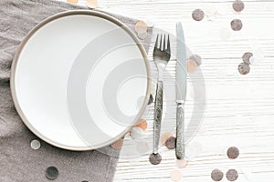 Stylish empty plate with vintage fork and knife on napkin on white table with confetti, flat lay. Modern set, serving for
