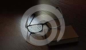 Stylish eyeglasses and book on wooden table in dark lighting