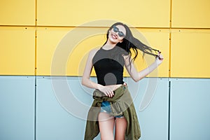 Stylish Fashion portrait of trendy cheerful casual young woman in black sunglasses posing near bright yellow urban wall. Beauty