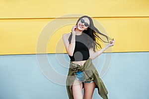 Stylish Fashion portrait of trendy cheerful casual young woman in black sunglasses posing near bright yellow urban wall. Beauty