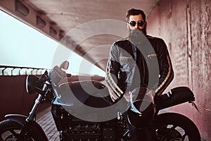 Stylish fashionable biker dressed in a black leather jacket with sunglasses holds a helmet sitting on his custom-made