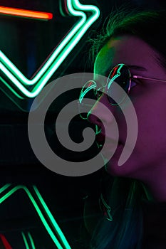 Stylish fashionable teenage girl in glasses with reflection looks up on the street with neon illumination of the city