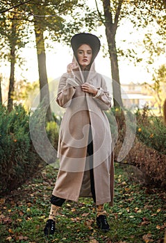 Stylish and fashionable young lady in beige coat and hooded cloak in a park on background of tree, beauty portrait of woman