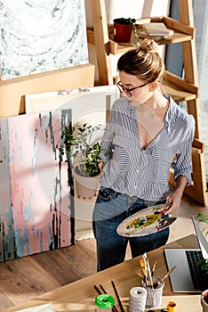 stylish female artist in eyeglasses holding plant and palette