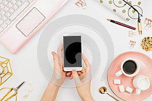 Stylish flatlay frame arrangement with woman`s hands holding white smartphone with black copyspace, pink laptop, coffee, planner