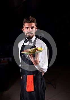 Stylish flunkey helpfully holds plate with prepared dish on a black background.