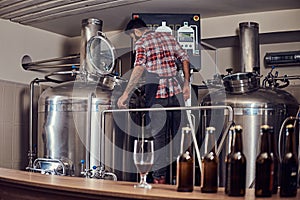 Stylish full bearded Indian man in a fleece shirt and apron controls the brewing process, standing near beer tank in the