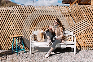 Stylish girl drinks coffee after shopping and cheking mail in social networks. Pretty young woman in brown coat and