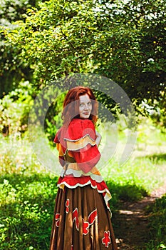 Stylish girl with freckles and red hair in red long dress holding red poppy flower at green field on nature