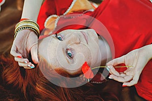 Stylish girl with freckles and red hair in red long dress holding red poppy flower at green field on nature