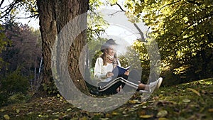 Stylish girl in the hat relaxing in the park, reading book while sitting on the grass.