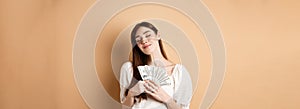 Stylish girl hugging money with pleased smile and closed eyes, like dollar bills, standing on beige background