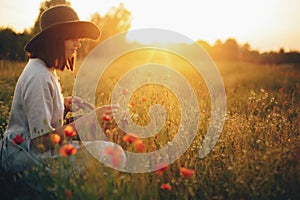 Stylish girl in linen dress gathering flowers in rustic straw basket, sitting in poppy meadow in sunset. Boho woman in hat