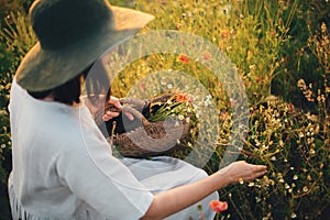 Stylish girl in linen dress gathering flowers in rustic straw basket, sitting in poppy meadow in sunset. Boho woman in hat