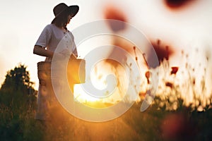 Stylish girl in linen dress gathering flowers in rustic straw basket, walking in poppy meadow in sunset. Boho woman in hat