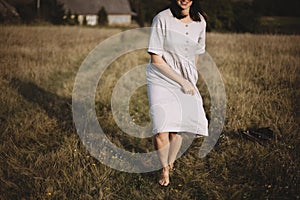 Stylish girl in linen dress and hat running barefoot in grass in sunny field at village. Boho woman relaxing in countryside,