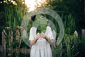 Stylish girl in linen dress holding big green leaf at face at wooden fence and grass. Portrait of boho woman in hat posing with