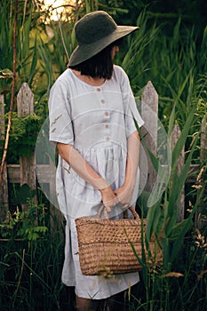 Stylish girl in linen dress holding rustic straw basket at wooden fence among green cane. Boho woman in hat relaxing and posing in