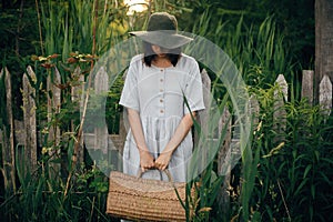 Stylish girl in linen dress holding rustic straw basket at wooden fence among green cane. Boho woman in hat relaxing and posing in