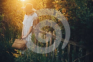 Stylish girl in linen dress holding rustic straw basket at wooden fence  in sunset light. Boho woman relaxing and posing in summer