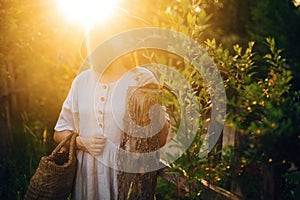 Stylish girl in linen dress holding rustic straw basket at wooden fence  in sunset light. Boho woman relaxing and posing in summer
