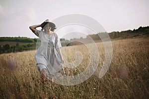 Stylish girl in linen dress running among herbs and wildflowers in sunny meadow in mountains. Boho woman relaxing in countryside,