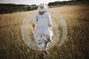 Stylish girl in linen dress running among herbs and wildflowers in sunny meadow in mountains. Boho woman relaxing in countryside,