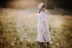 Stylish girl in linen dress walking among wildflowers in sunny meadow in mountains. Boho woman relaxing in countryside flowers at