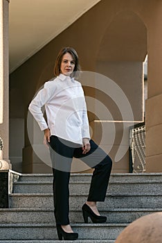 Stylish girl is photographed on steps of beautiful building. Decisive woman is dressed in classic white shirt and black pants